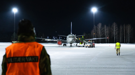 The photo shows an airport at night with an aircraft being assisted by ground personnel. Two people in reflective vests, one of whom has "MILITARY GUARD" written on the back, stand in front with their backs to the camera. The aircraft, illuminated by powerful lights, is surrounded by snow-covered ground and dark trees in the background. A tow truck is visible next to the aircraft, and there is activity around the plane in preparation for departure or after arrival.