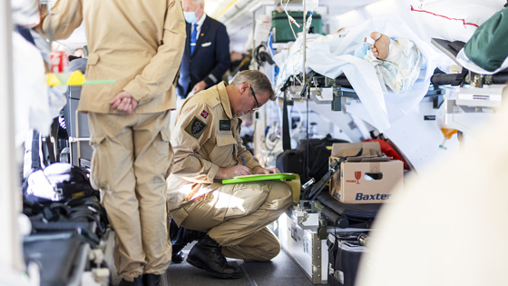 The photo shows a man in a beige military uniform crouching in an aircraft and writing on a green clipboard. The background reveals a range of medical equipment and a patient lying on a stretcher, covered with white sheets. Another person in uniform stands in the background, and there are several boxes and pieces of equipment visible around the cabin.