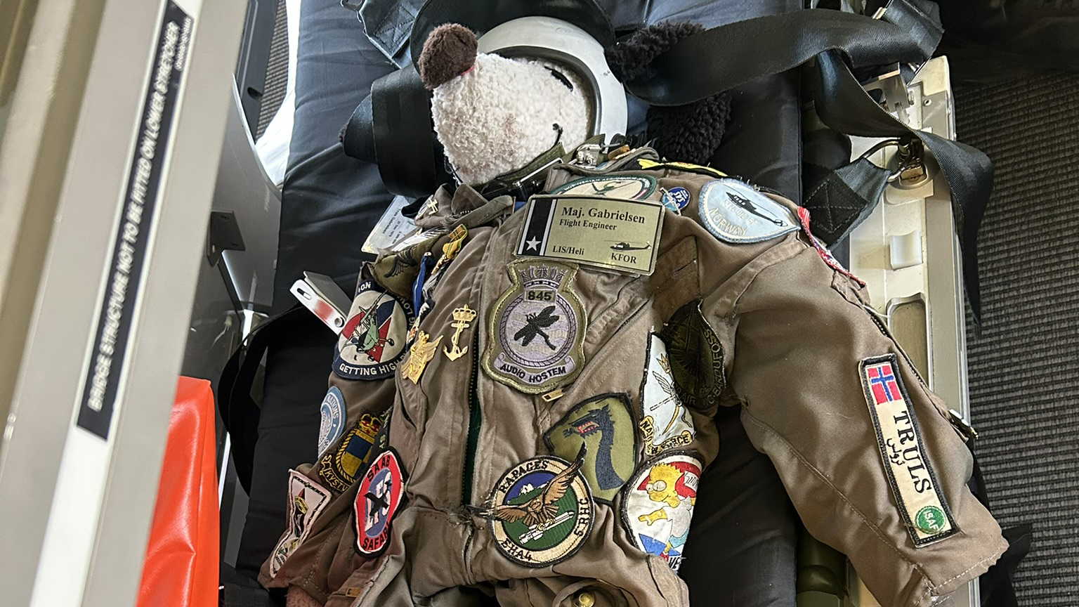 The photo shows a well-worn military jacket adorned with various patches and insignias from different military units and operations. The jacket is decorated with patches representing different nations and special units, including a patch that says "Maj. Gabrielsen, Flight Engineer." A teddy bear with pilot goggles is also attached to the jacket, adding a personal and humorous touch to the otherwise serious military gear. The jacket is placed in an aircraft seat, suggesting that the owner is an active or former military aviator.