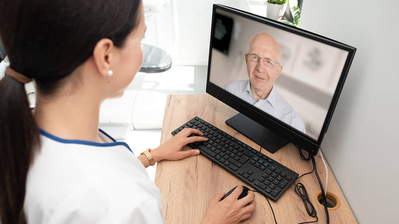 Bildet viser en kvinnelig lege i en hvit uniform som sitter ved et skrivebord og bruker en datamaskin. Hun ser på en skjerm hvor en annen person er synlig i et videomøte. Kvinnen bruker tastaturet og musen, og det ser ut til at hun er engasjert i en digital samtale eller konsultasjon. Kontormiljøet er lyst og moderne, med en plante og noen dekorative elementer i bakgrunnen.